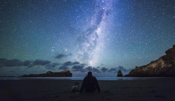 A person is sitting at a beach facing the water under a bright starry night sky with blue and purple hues. 