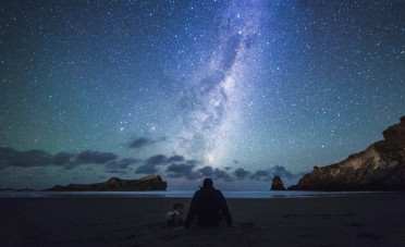 A person is sitting at a beach facing the water under a bright starry night sky with blue and purple hues. 