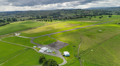 An aerial photograph showing a rural airfield with a single runway, adjacent buildings, and large expanses of green farmland stretching toward distant hills beneath a cloudy sky