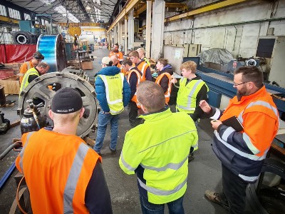 A group of students in high-vis gear are observing engineering work in a warehouse setting.