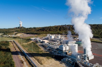 A geothermal cloud is rising from a large work site. In the background there is blue sky and green landscape.