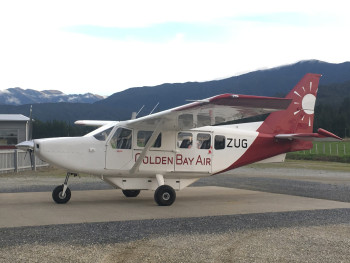 A white and red Golden Bay Air aircraft sits on an airstrip with mountains and cloudy skies in the background.