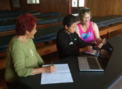 Two women help a boy with his homework using digital technology such as a laptop, tablet and mobile phone