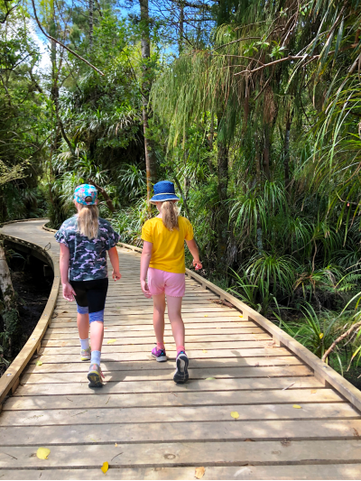 In an outdoor nature setting, two young people are walking alongside one another on a wooden plank pathway.