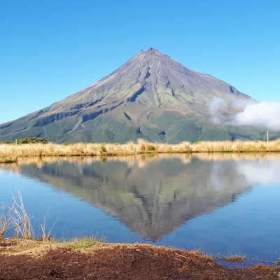 Mount Taranaki with a vibrant blue sky above, dried grass in front and the mountain's reflection on water in the foreground.