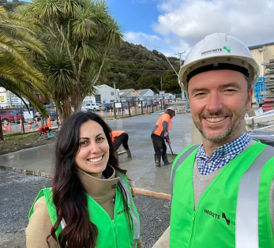 Two people smiling wearing green high-visibility vests in front of a concrete work site.