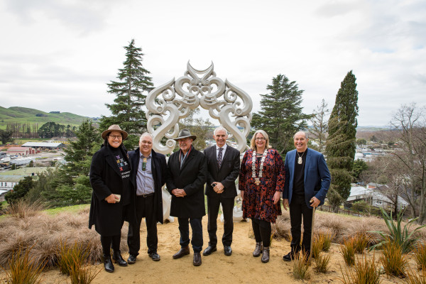 Full body shot of six people standing in a line, dressed professionally, smiling at the camera. They stand in front of a scenic background with houses, trees and hills and a grey sky above them.