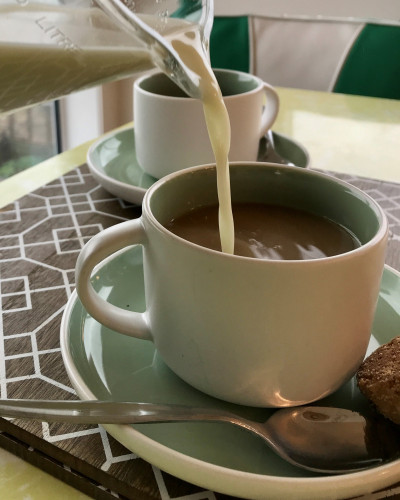 Glass jug of oat milk being poured into a white and green mug filled with tea.