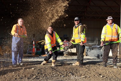 A group of four holding shovels and wearing high-vis gear are standing together. One has just launched some dirt in the air.