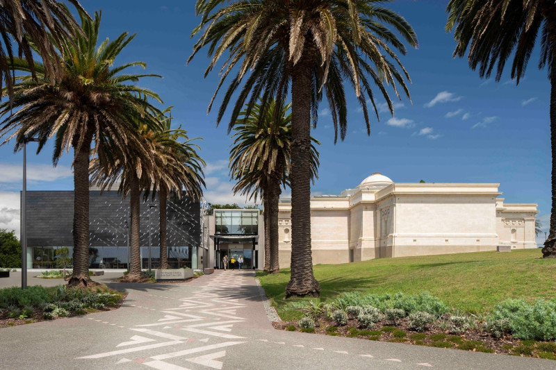 A pathway featuring grey concrete and white triangular patterns and flanked by large palm trees, leads to a gallery building entrance.