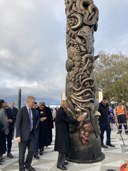 A tall piece of Māori art with carvings of sea creatures on it in a town centre. Lots of people around it dressed in business-wear, including Prime Minister Chris Hipkins.