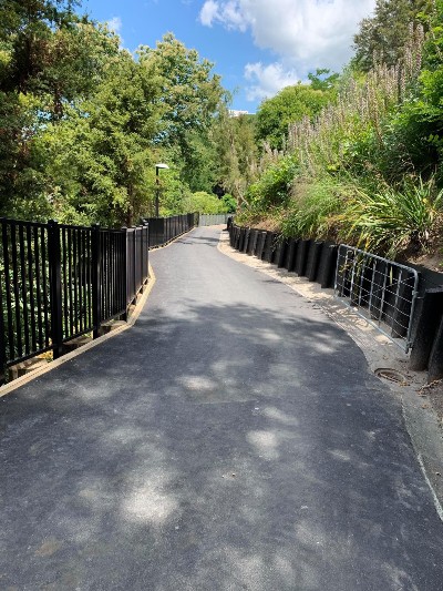 A view down a sloping paved pathway surrounded by fence, trees and other greenery.