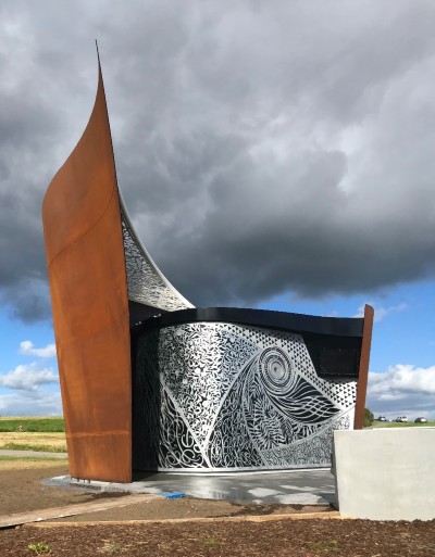 A large outdoor sculpture that features a copper and silver panels. The silver panels have highly detailed patterns cut out of them.