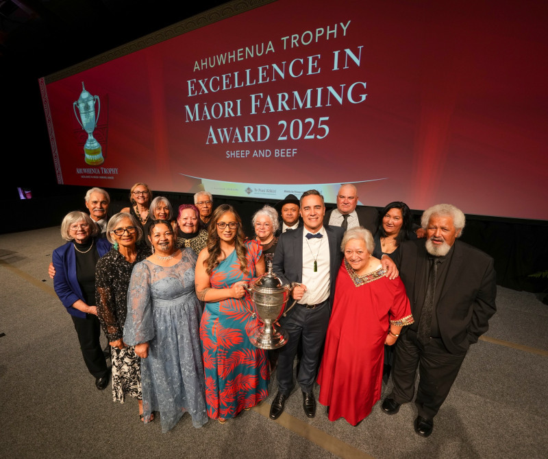 A group of happy people in formal attire stand together surrounding a large trophy being held by a woman in a blue dress with tropical red leaves print.