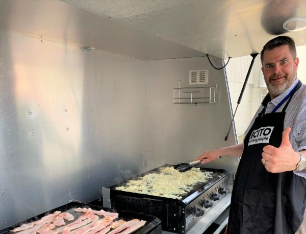 A person wearing a black BCITO apron is doing the thumbs up sign while grilling bacon and onions at a barbecue.