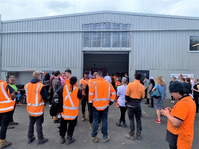 People gathered outside an industrial building; many of them are wearing orange high-vis gear.