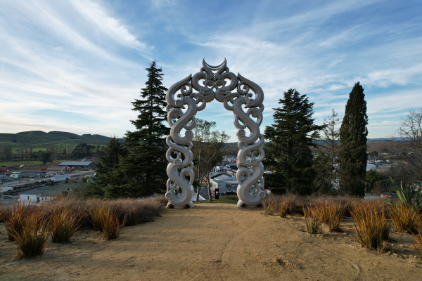 A white archway made of wooden Māori carvings on a clay dirt ground with small plants either side of it, and trees behind.