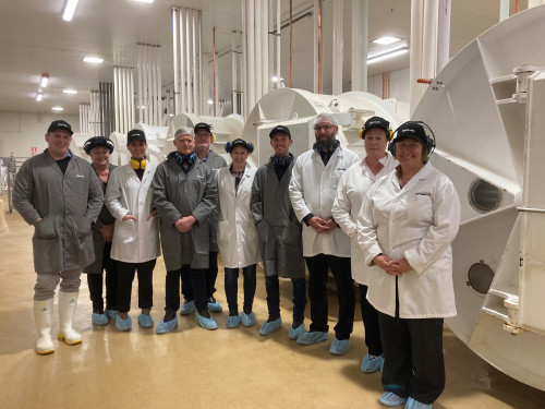 Ten people stand in a clinical lab environment, wearing lab coats, hats, earmuffs, and covers over their shoes.