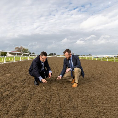 Two men wearing business suits kneel down, touching the dirt on the ground of a newly opened racecourse.
