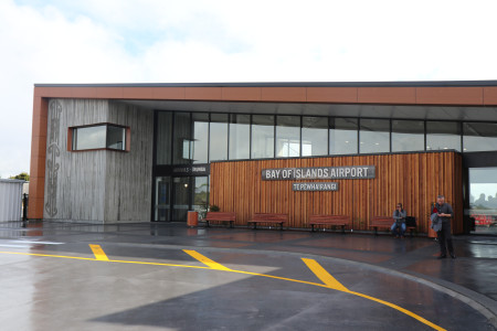 The outside of the Bay of Islands Airport - wooden panels on the outside, Māori designs and a sign saying 'BAY OF ISLANDS AIRPORT'. Two people are waiting outside.
