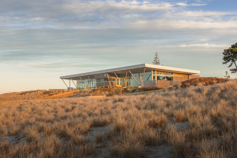A new single storey building with a flat roof and windows all around it, sits atop sand dunes