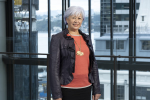 Mid shot of a woman with short, white hair, standing, wearing a red shirt, black leather blazer and gold necklace.