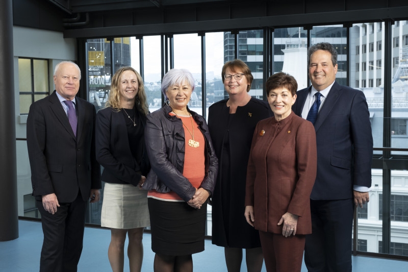 Group photo of six of the board members, showing from their knees upwards, all dressed professionally in a building with glass windows looking out to a city view.