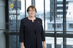 Mid shot of woman standing, with short dark hair, wearing a navy blue dress and blazer, glasses and a gold broche.