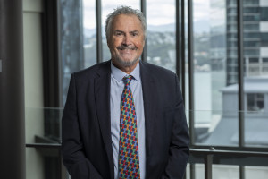 Man in a business suit with short grey hair and facial hair stands in front of a window in a high rise office building