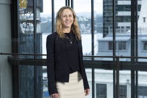 Mid shot of woman with shoulder-length blonde, curly hair, wearing a black blazer and shirt, and white skirt.