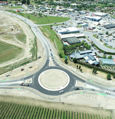 Bird's eye view of a newly constructed roundabout with a big dirt patch in the center. Cars are going around it. There is an industrial area along one side of it and grass fields on the other side