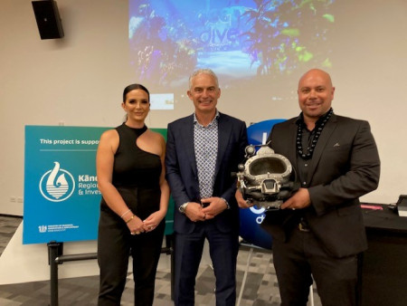 Three people dressed formally, stand in a room in front of a Kānoa sign. Man on right holds an underwater diving mask.