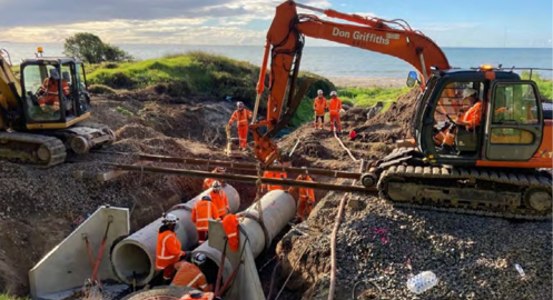 Diggers and cinstruction workers wearing hi-vis installing large concrete pipe in a trench, going out to sea