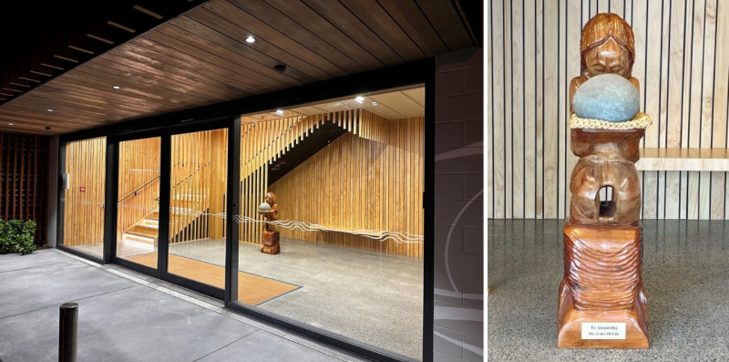 Looking through glass inside a new building with wooden panels on the walls, wooden stairs and a Māori carving