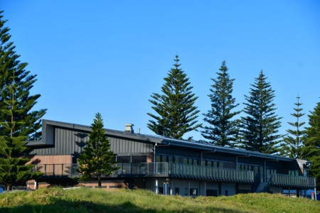 A modern, two storey building with grass in front of it and pine trees all around. It has a slanted roof