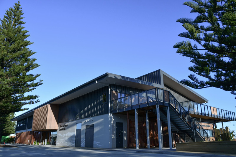 A new two storey building, coloured grey and black. A blue sky above and tall pine trees stand either side of it
