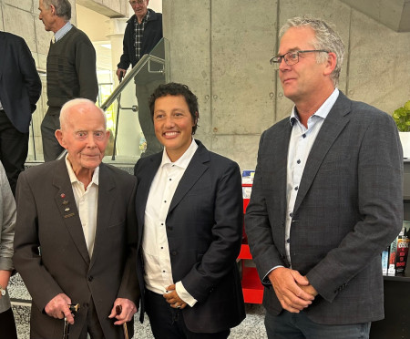 An elderly man and a middle aged woman and man stand together, all dressed formally, smiling. Inside the new community centre.