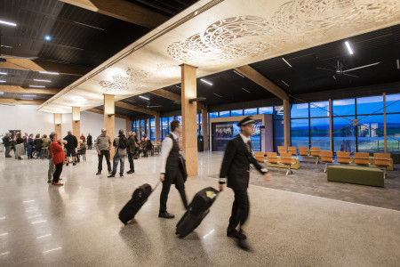 A modern airport terminal, blurry pilots rolling suitcases walk past, passengers and members of the public waiting in the background.