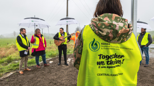 A close up shot of the back of a yellow hi vis vest which has the Kānoa-RDU logo, and the words 'Together we thrive! E ora Ngātahi ana' and 'CENTRAL HAWKE'S BAY'. Four other people are standing in the background wearing the vests and holding umbrellas, standing in front of a grass paddock.