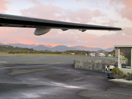 Image taken out of a plane window. The wing of the plane goes across the top of the image and pink and orange colours are on the hills in the distance from the sky. The plane is still on the ground.