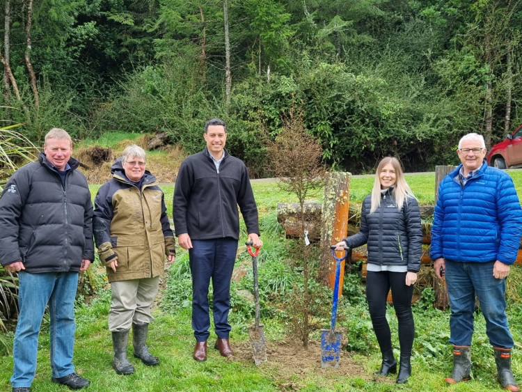 Five people stand on grass, in front of a dense bush area, next to a freshly planted tree, which is the height of a person. All people are wearing gumboots and rain jackets, and are standing in a line, smiling at the camera.