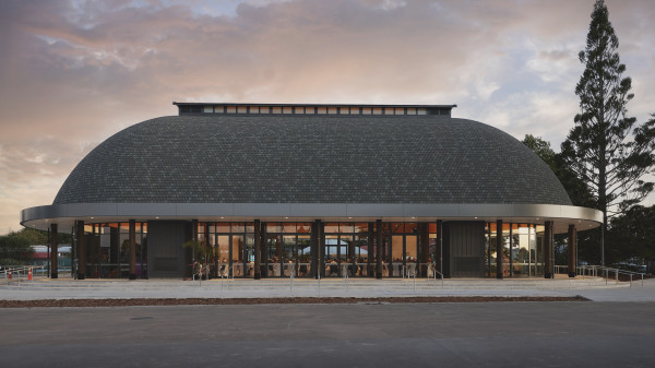 A new building with a rounded brick rood. There are lights on inside and people sitting at tables. Lots of glass windows and doors around the outside of the building