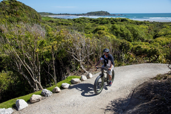High angle shot of a person riding a mountain bike on a gravel path with trees on either side of it and the ocean in the background. A sunny day. The cyclist is wearing a helmet and casual clothes.