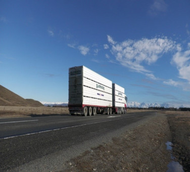 Low angle shot of a truck driving along a road in a remote location. There's a blue sky above and snowy mountains in the distance