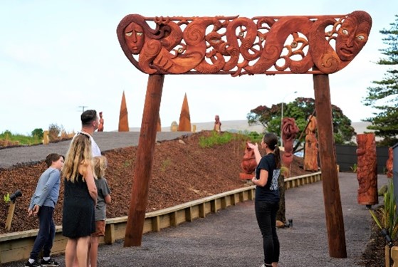 A man and his children listen to a person talk about a wooden archway which has traditional Māori designs carved into the top of it. Other wooden carvings are seen in the background further along the walkway