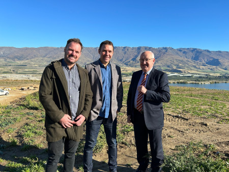 Three men dressed in businesswear stand on a field of grass with hills and a vibrant blue sky in the background.