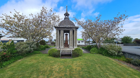 A concrete structure with pillars, a domed roof and a point on top, sitting on a lawn with trees either side