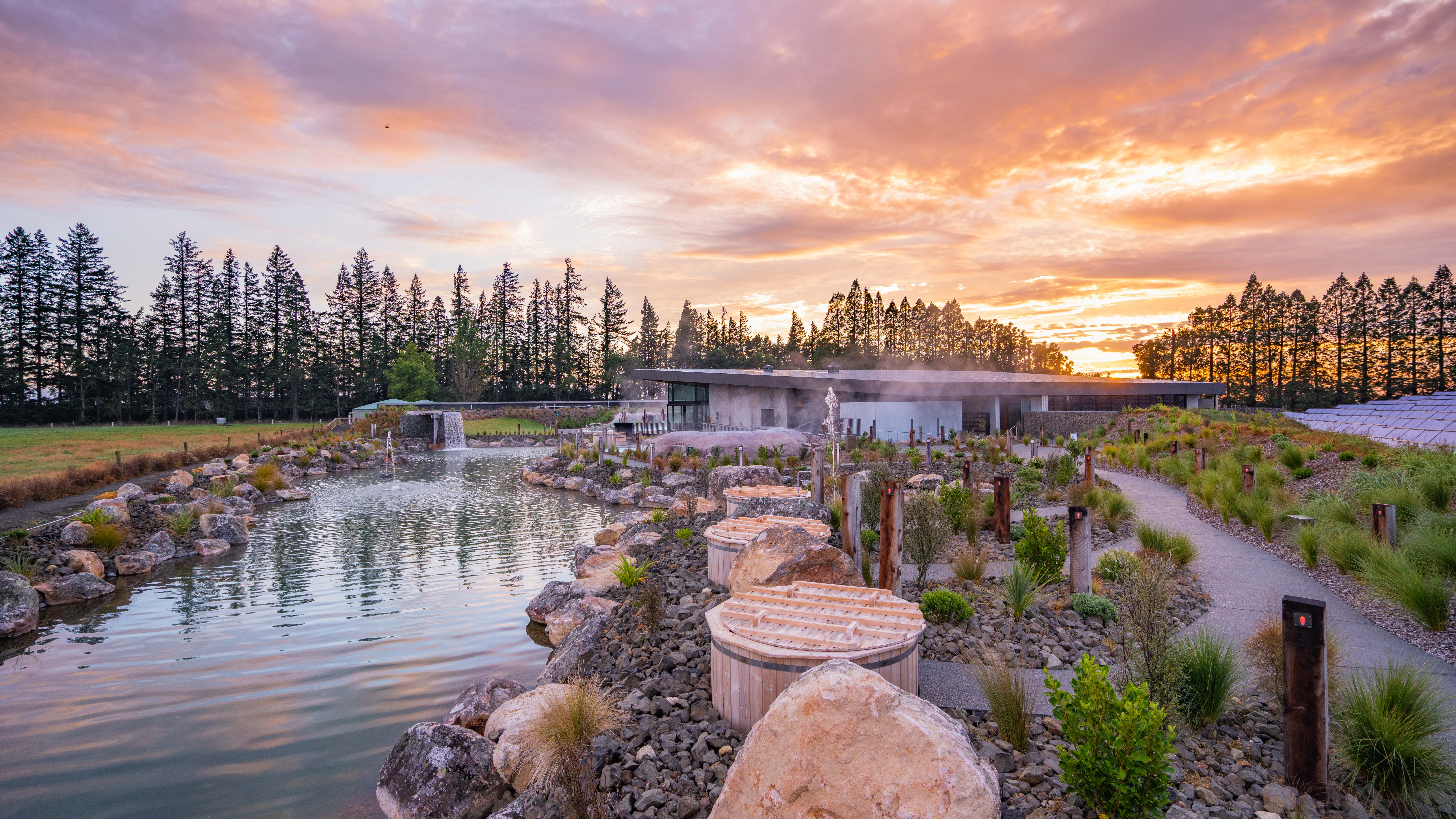 Outdoor spa pools alongside rocks, water and new plants, with a building and trees in the background