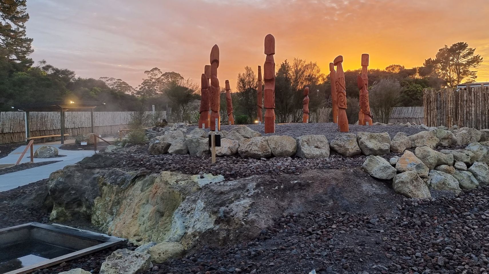 Māori carved figures standing in a circle outside, with rocks and steam from natural hot springs surrounding