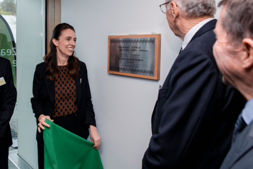 The Prime Minister holds a piece of green fabric, and is looking at a silver plaque with a wooden frame on a white wall. Other people are in the edge of the photo smiling, looking at the plaque and the Prime Minister.